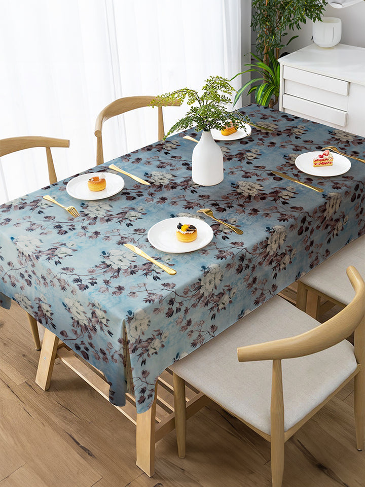 Dining table set with a floral tablecloth, plates, and cutlery in a bright room.