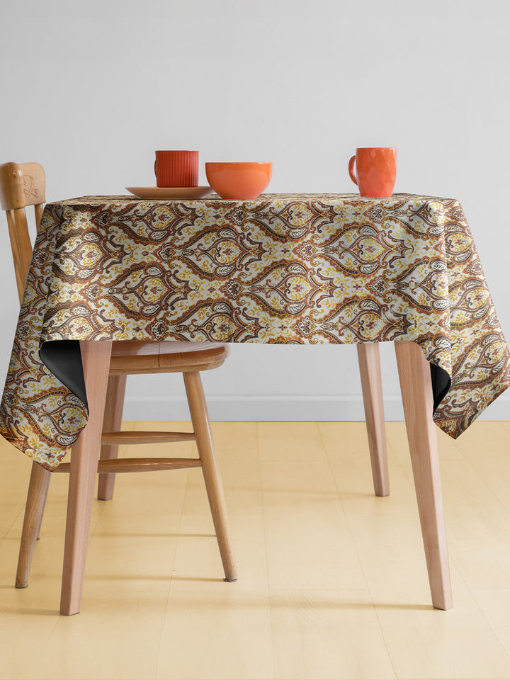 Table with a patterned tablecloth, wooden chairs, and orange cups on a light background