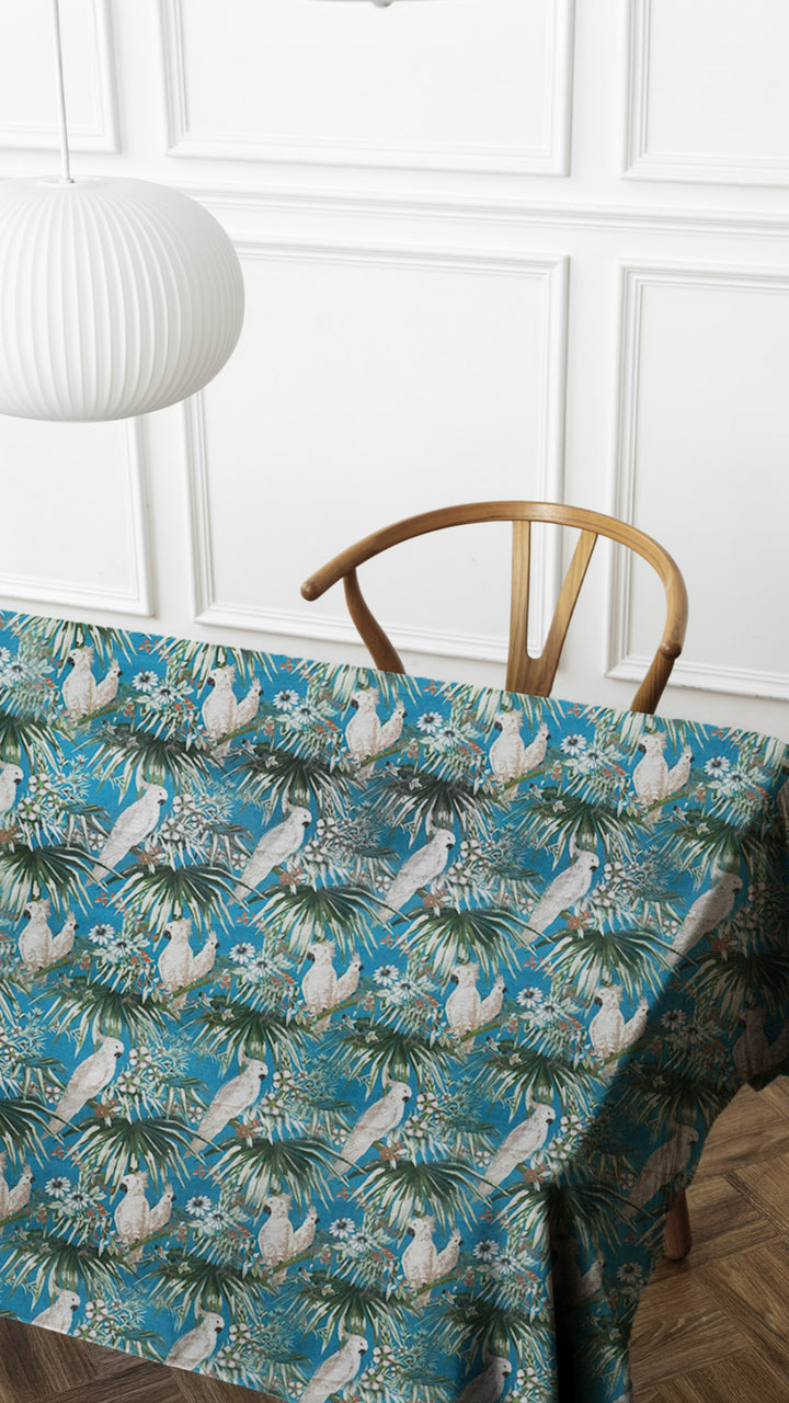 Tablecloth with parrot and palm tree pattern on a table with a wooden chair against a white paneled wall.