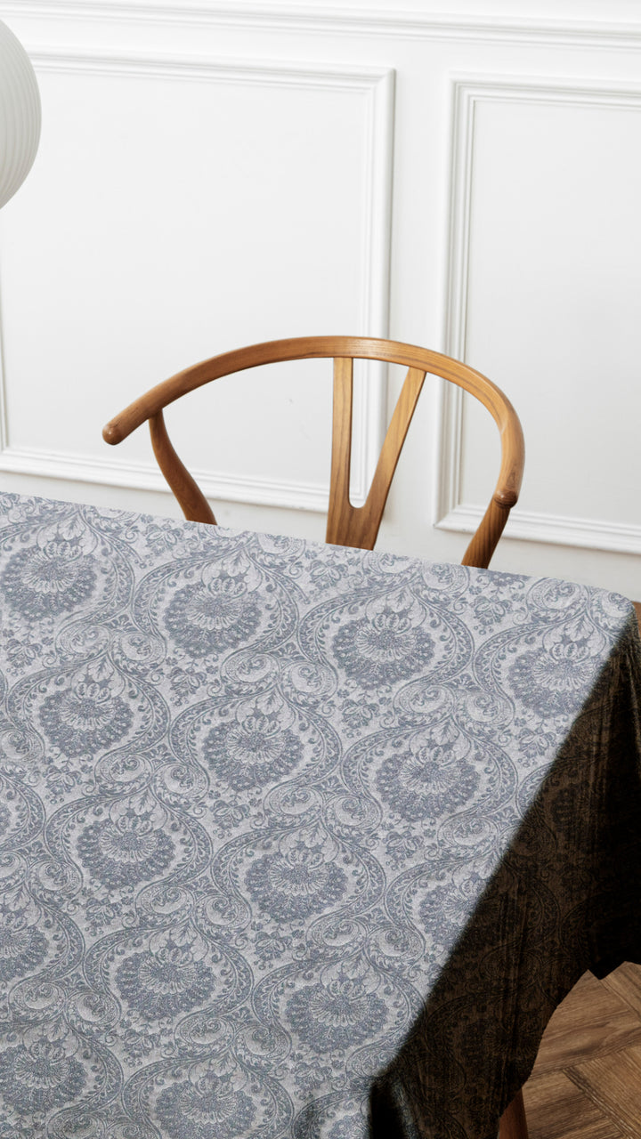Dining table with a patterned gray tablecloth and wooden chair against a white paneled wall.