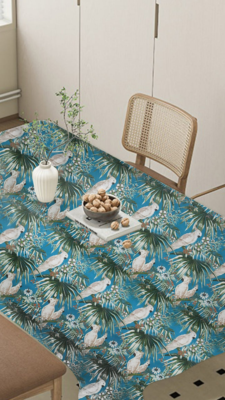 Table with a blue floral tablecloth in a room with a chair and vase.
